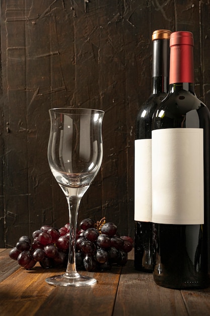 Empty wine glass next to two bottles of red wine on a rustic wooden table and dark background. Bunch of grapes behind.