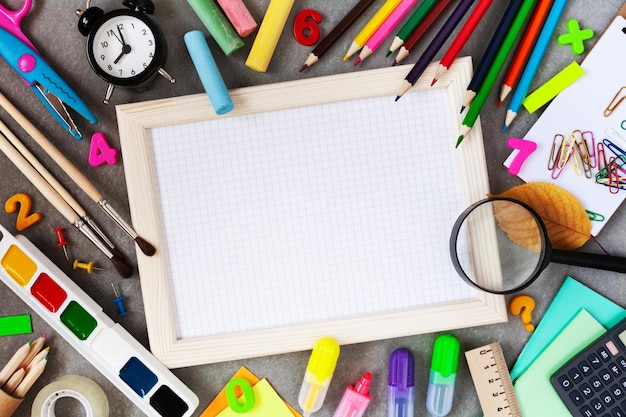 Empty wooden frame with squared paper alarm clock different stationery and colorful supplies on gr