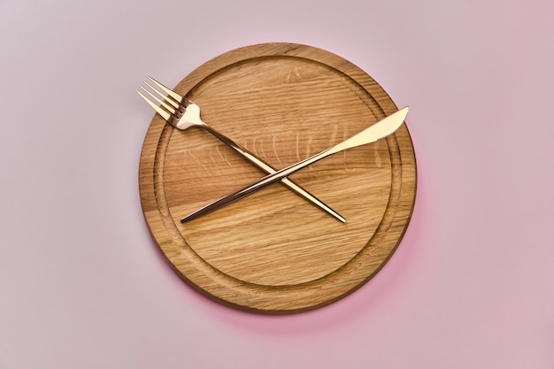 Empty wooden round tray or trencher with cutlery as clock hands on pink surface