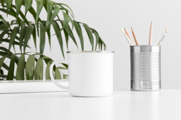 Enamel mug mockup with workspace accessories on a white table and a palm plant