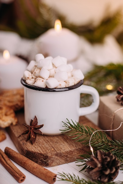 Tazza smaltata di cioccolata calda o caffè con marshmallow e biscotti. attorno ai rami degli alberi, regali e candele accese. atmosfera natalizia. cartolina o sfondo invernale.