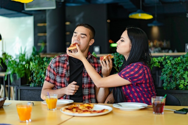 Enamored woman gives a guy a piece of pizza in the restaurant on the background of the interior of green plants Young couple have lunch at a cafe