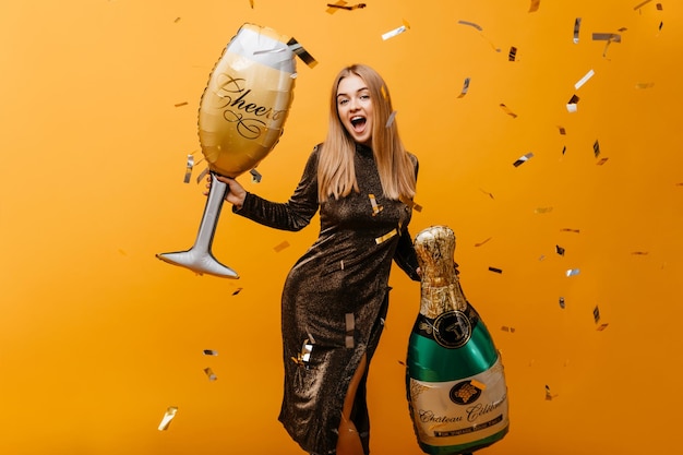 Incantevole ragazza di buon umore in posa con una grande bottiglia di champagne giovane donna divertente in abito scintillante che tiene un bicchiere di vino