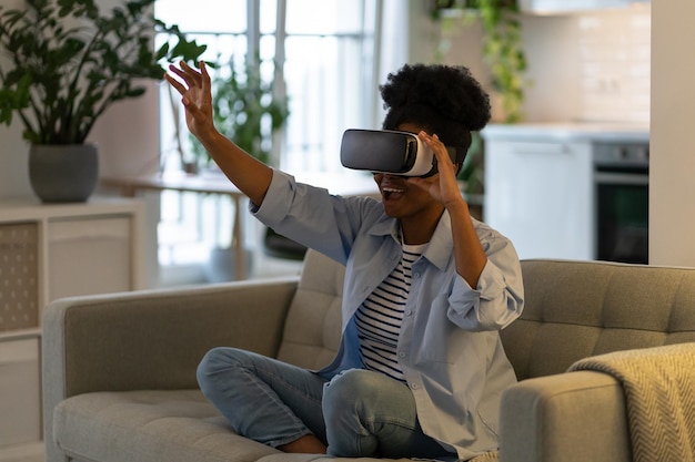 Encouraged African American woman using virtual reality glasses to watch 3D movies sits on sofa