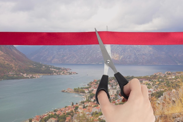 End of quarantine covid19 the beginning of travel the opening\
of borders sale of tour packages a hand cuts a red ribbon with\
scissors overlooking the kotor bay of the adriatic sea in\
montenegro