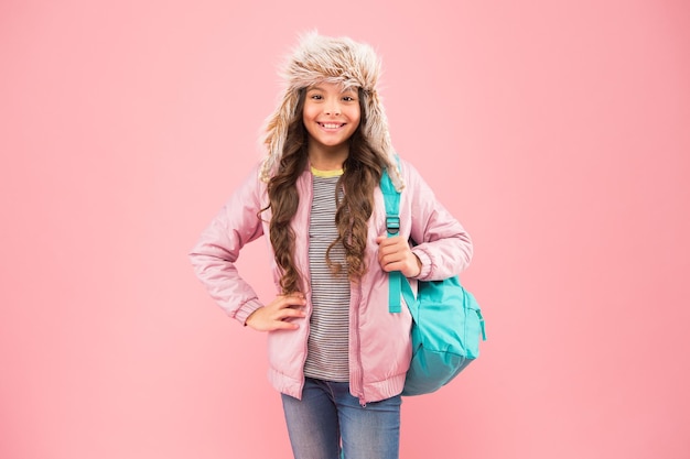 The end of school year. no more exams. small happy girl back to school. winter holiday and vacation. kid warm clothes pink background. happy child earflap hat. schoolgirl with backpack go home.