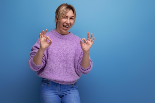 Energetic cheerful blondie young woman in a lilac sweater with a kind heart on a bright background