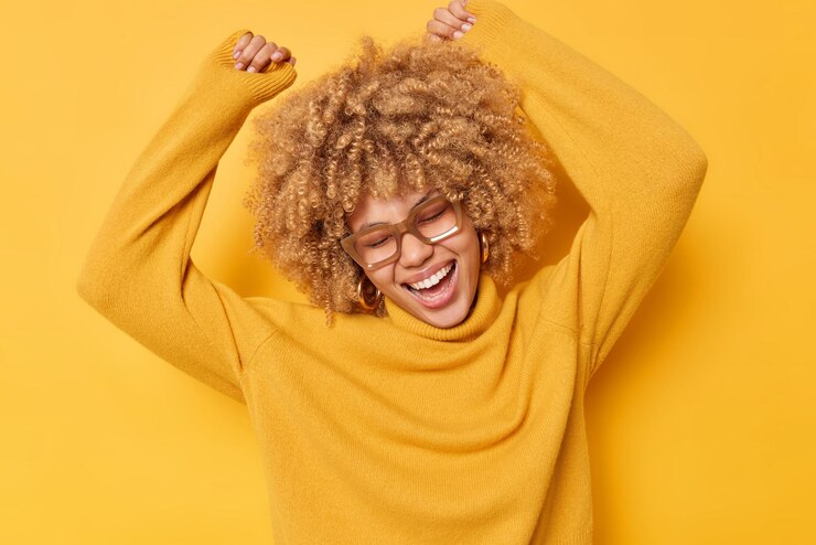 Energetic curly haired young woman dances carefree keeps arms raised feels joyful celebrates victory wears transparent glasses and sweater isolated over vivid yellow background feels like winner