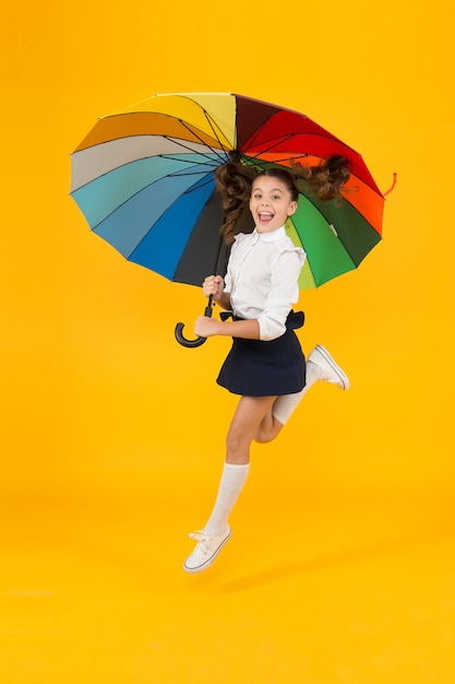 Energetic look. Adorable girl jumping with autumn look on yellow background. Cute little schoolchild having glamour look with colorful umbrella. School look of small child.