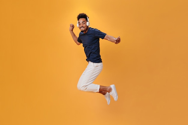 Energetic man in stylish outfit jumping on orange background and listening to music on headphones Cool guy un blue shirt having fun on isolated backdrop