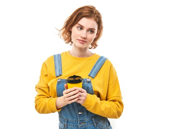 Energetic redhead girl holding and drinking yellow cup of takeaway coffee smiling and enjoying drink isolated on white background