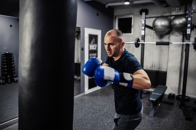 Energie- en conceptboksen. Een toegewijde man draagt bokshandschoenen en sportkleding in een overdekte moderne fitnessruimte met apparatuur. De mens in sport en boksen maakt zich net klaar om een grote zwarte bokszak te raken