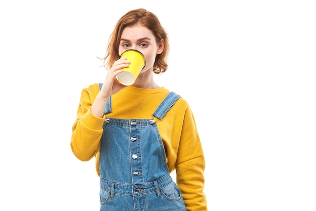 Energieke roodharige meid die een gele kop afhaalkoffie vasthoudt en drinkt, glimlachend en genietend van een drankje geïsoleerd op een witte achtergrond