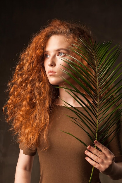 에너자이저는 녹색 열대 식물 야자나무 가지가 있는 아름다운 젊은 곱슬 생강 머리 여성이 어두운 벽 스튜디오 복사 공간 배경에 포즈를 취했습니다.
