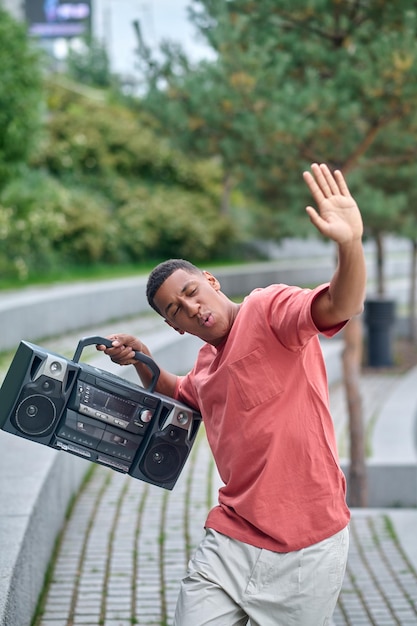 Energy of life. Dark-skinned energetic guy with closed eyes holding record-player raising hand in dance move in park during day