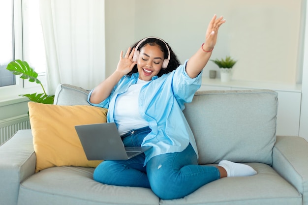 Energy mood Happy black body positive lady in wireless headphones listening music online on laptop sitting on sofa