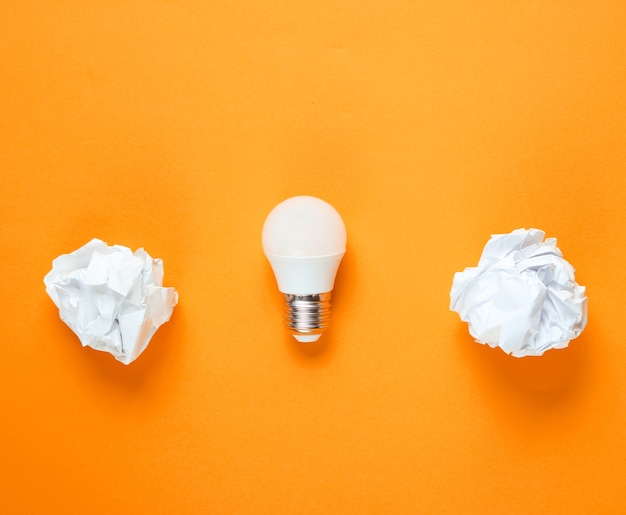 Energy saving light bulb and crumpled paper balls on orange background. Minimalistic business concept, idea. Top view