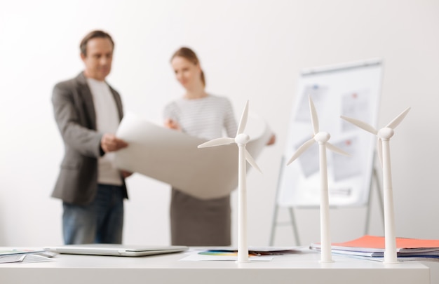 Energy saving. Selective focus of wind turbines standing on the table while professional colleagues engineers discussing eco project and holding a blueprint