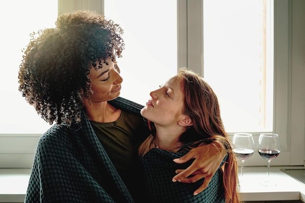 Photo engaged couple of young lesbian women holding together against the kitchen window drinking red wine