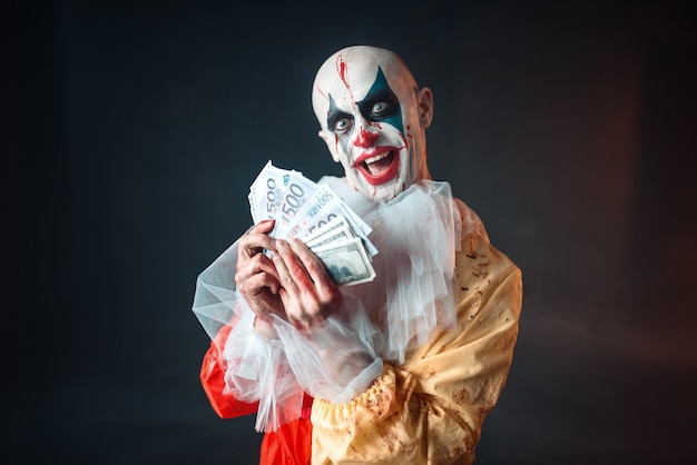 Enge bloedige clown met gekke ogen houdt fan van geld. Man met make-up in carnavalskostuum, gekke maniak