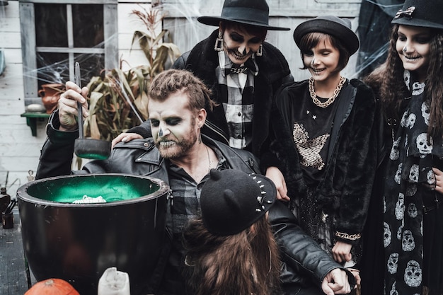 Enge familie moeder vader dochters vieren halloween Ketel van toverdrank Angstaanjagende zwarte schedel halfgezicht make-up heks kostuums stijlvolle afbeeldingenHorrorpret op kinderfeestje in schuur op straat