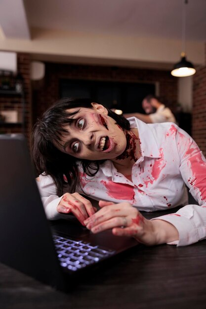 Enge, gruwelijke zombie die laptop gebruikt aan het bureau, ondode lijk die probeert te werken op de computer in het opstartkantoor. Griezelig agressief hersenetend monster dat er angstaanjagend en vreselijk uitziet, sinister gevaar.