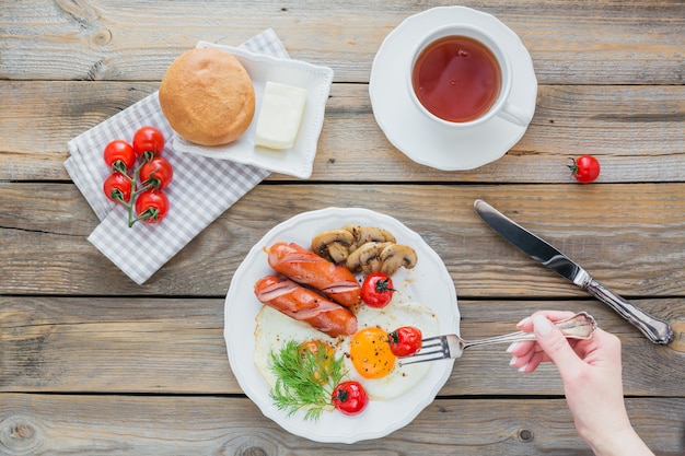 Engels ontbijt met gebakken eieren, worstjes, champignons, gegrilde tomaten en kopje thee op rustieke houten tafel. bovenaanzicht