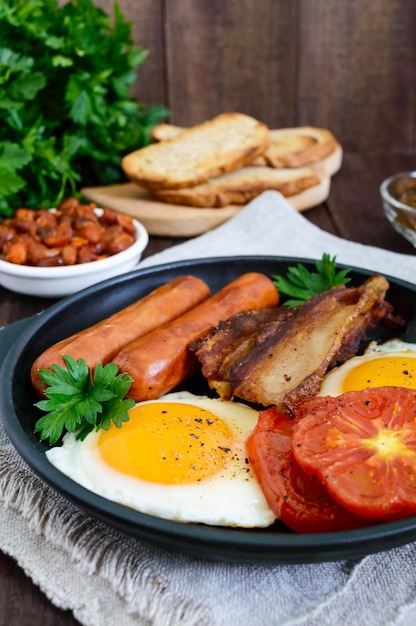 Engels ontbijt: worstjes, spek, tomaten, ei, bonen in saus, gebakken champignons, toast op een donkere houten tafel. Verticale weergave. Traditionele klassieke gerechten uit Engeland.