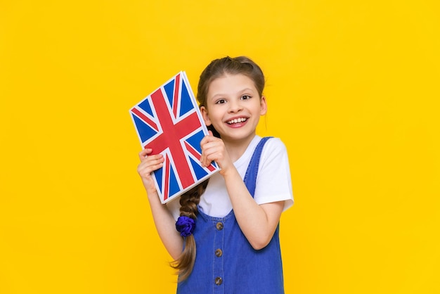 Engels voor kinderen Een klein meisje heeft een leerboek met een Engelse vlag in haar handen Taalonderwijs voor kinderen Gele geïsoleerde achtergrond