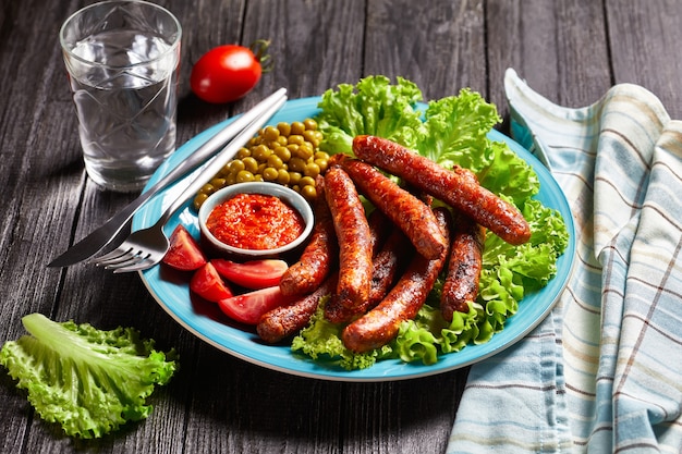 Engelse maaltijd: varkensworst chipolata geroosterd en geserveerd op een blauw bord met tomatenketchup, tomaten, groene sla en erwten op een rustieke houten ondergrond, bovenaanzicht, close-up