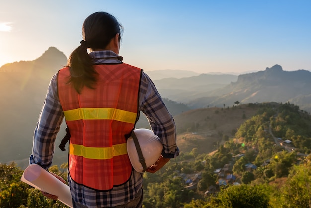 Engineer Asian man a white helmet and blueprint, looking at the mountain In which there is a village built on the top of the hill