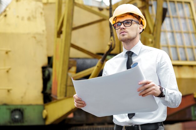 Инженер-строитель в каске держит чертежи на строительной площадке