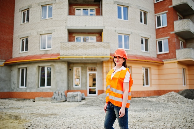 Engineer builder woman in uniform waistcoat and orange protective helmet against new building