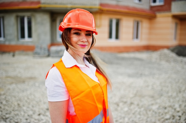 Engineer builder woman in uniform waistcoat and orange protective helmet against new building