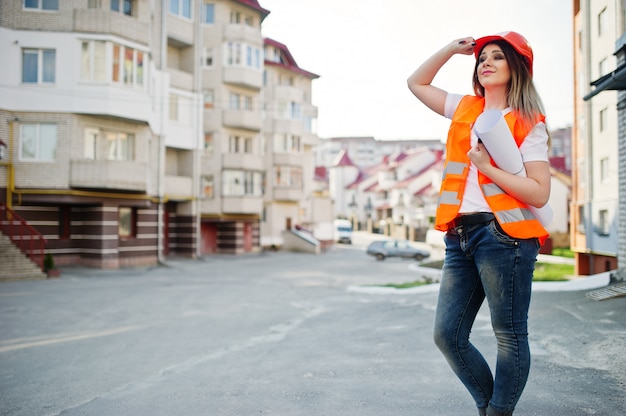 制服チョッキとオレンジ色の保護ヘルメットのエンジニアビルダーの女性は、新しい建物に対してビジネス描画紙ロールを保持します。