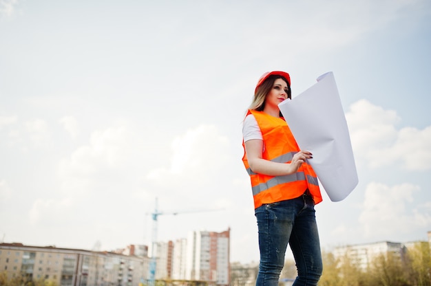 Engineer builder woman in uniform waistcoat and orange protective helmet hold business paper against new buildings with crane. 