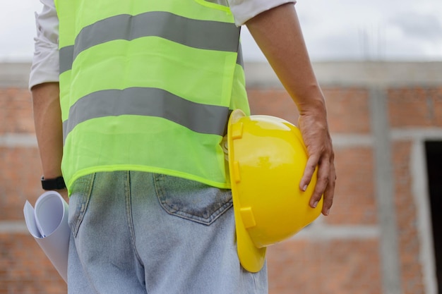 Engineer concept the male constructing staff that wears a white\
top covered with bright lime vest, a black watch, and light blue\
jeans standing and holding a mustard safety hat.