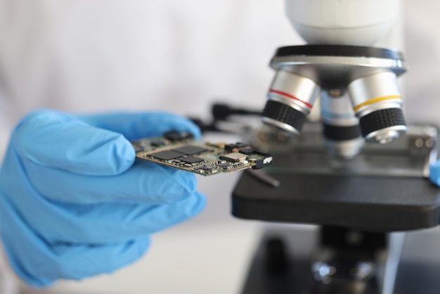 Engineer in glove holds microcircuit against background of microscope