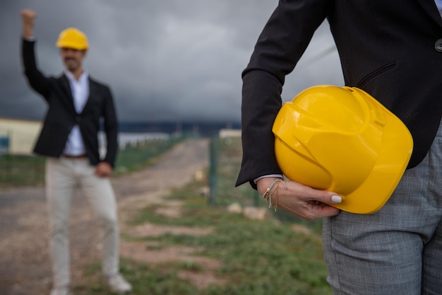 An engineer holds a hardhat in one hand while her colleague behind celebrates a success at work