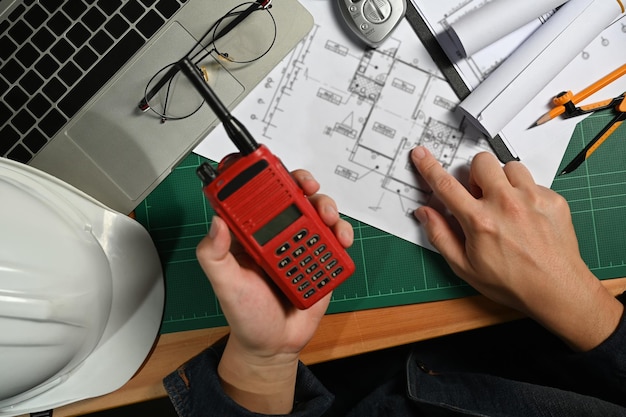 Engineer man hand holding walkie talkie working on blueprint at wooden office desk Above view