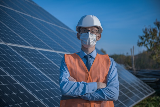 엔지니어, 유니폼 및 마스크, 헬멧 안경 및 태양 광 발전소에서 태양 전지 패널의 배경에 작업 재킷 남자. 기술자가 유지 보수를 확인합니다.