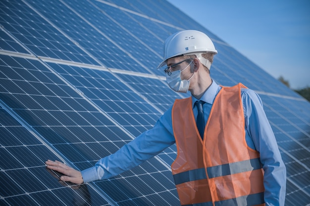 Engineer, man in uniform and mask, helmet glasses and work jacket on a of solar panels at solar station