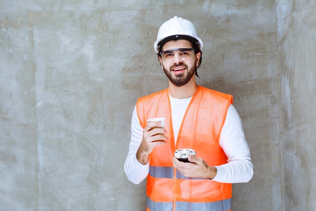 Uomo dell'ingegnere in casco bianco e occhiali protettivi che tengono una sveglia e una tazza di bevanda.
