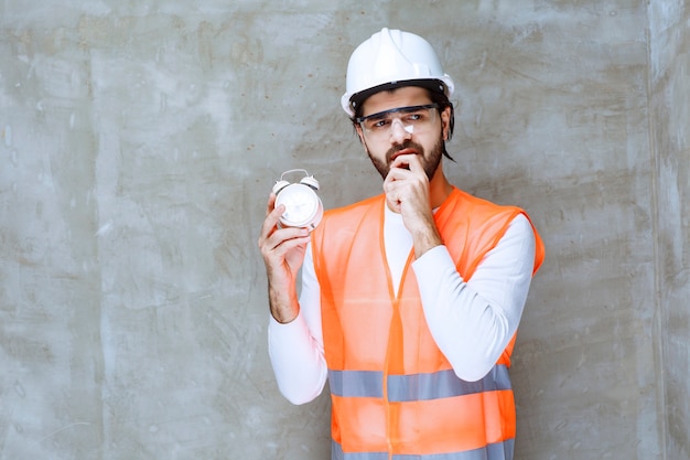 Engineer man in white helmet and protective eyeglasses holding an alarm clock and looks confused and lost.