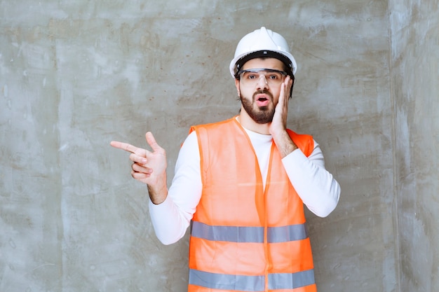 Engineer man in white helmet and protective eyeglasses pointing at his colleague or at something aside.