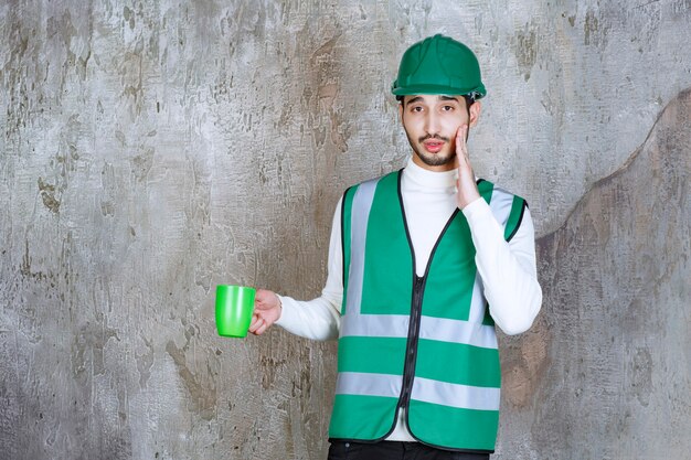 Engineer man in yellow uniform and helmet holding a green coffee mug and looks surprized.