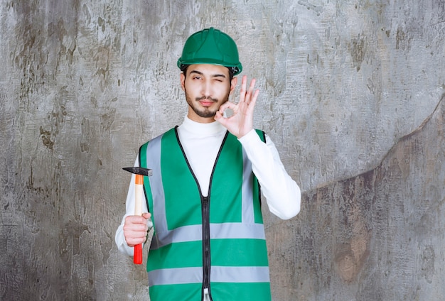 Foto ingegnere in uniforme gialla e casco che tiene un'ascia con manico in legno per la riparazione e mostra il segno della mano di piacere