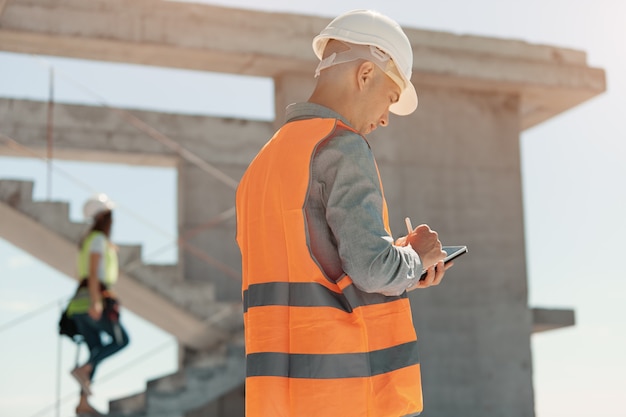 Un ingegnere con un giubbotto arancione e un casco bianco di controllo della costruzione conduce un'ispezione con un tablet in mano.