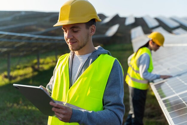 Engineer of solar power plant blogging on company page on\
social network young man in yellow vest holding tablet in hands\
posting new content on background of solar panels