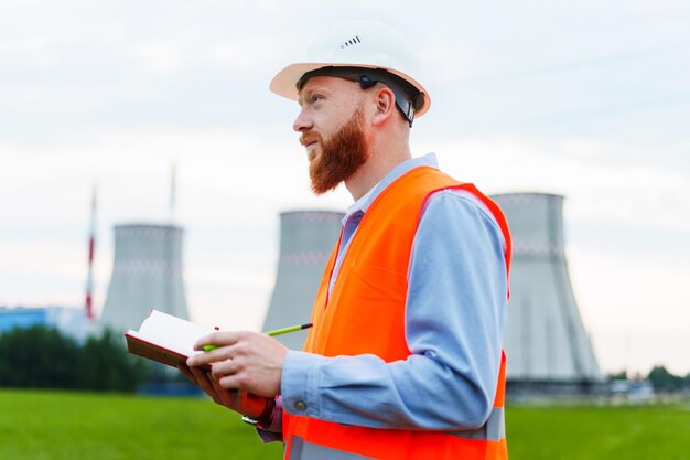 Photo an engineer with a notebook on the background of a power plant the man is wearing a white helmet and an orange vest